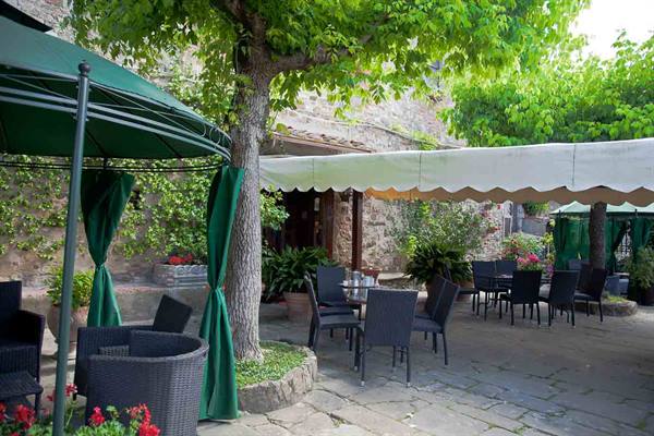 Empty tables under green umbrellas and awnings, stone courtyard shaded by trees