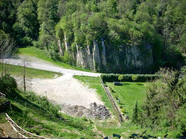 Dirt road and green meadow in front of a rocky cliff covered with trees