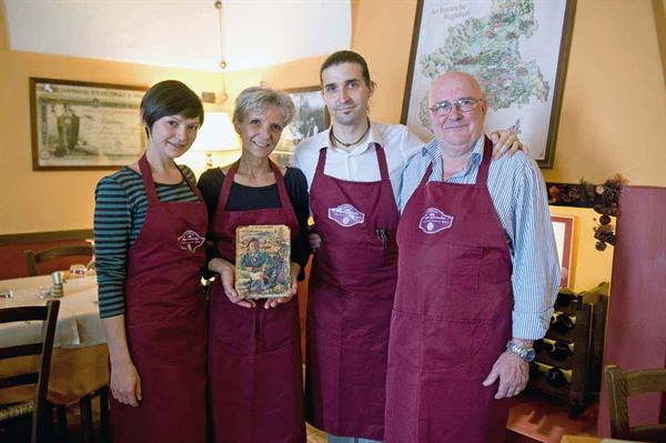 Four people in burgundy aprons pose in a trattoria, one holding a book