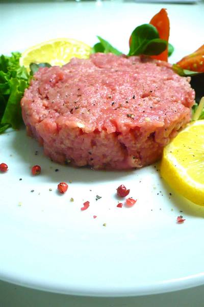 Raw beef tartare on white plate with lemon, lettuce and pink pepper