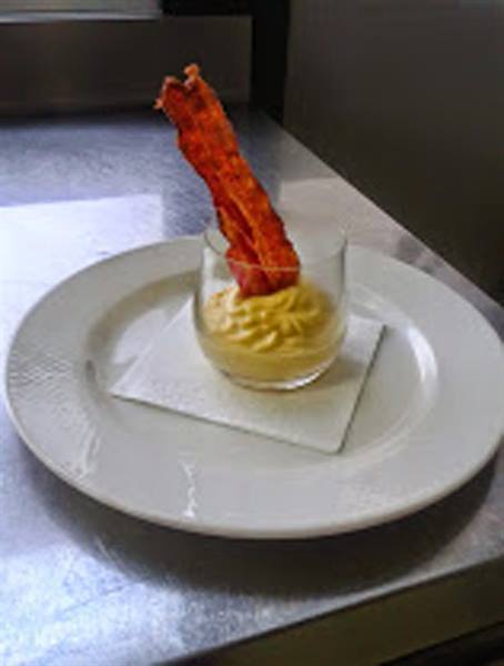 Glass cup with yellow cream and crispy bacon strip, on white plate