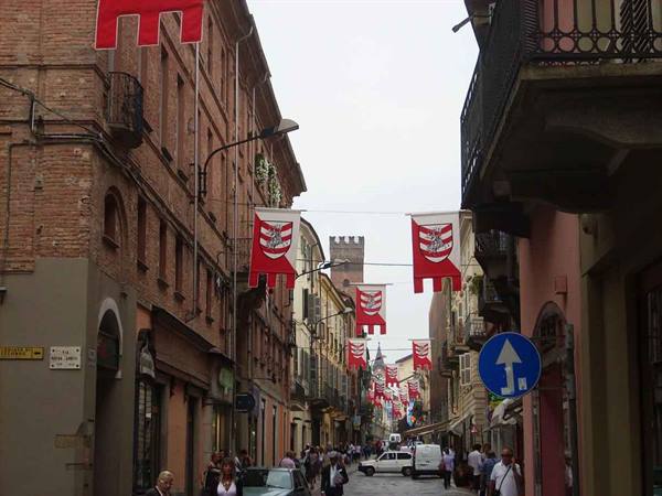 Street with red and white flags hung between brick buildings, pedestrians and cars