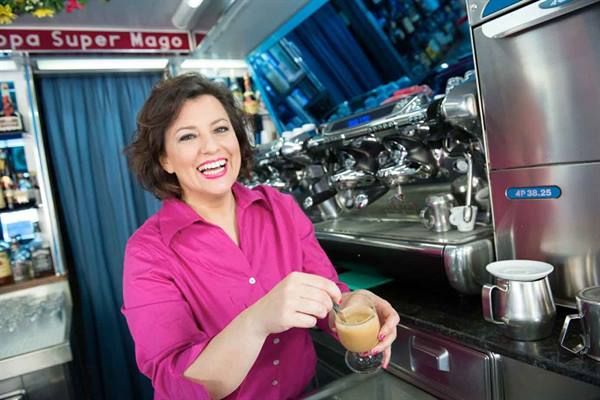 Smiling woman in fuchsia shirt stirring a coffee at the counter, chrome espresso machine