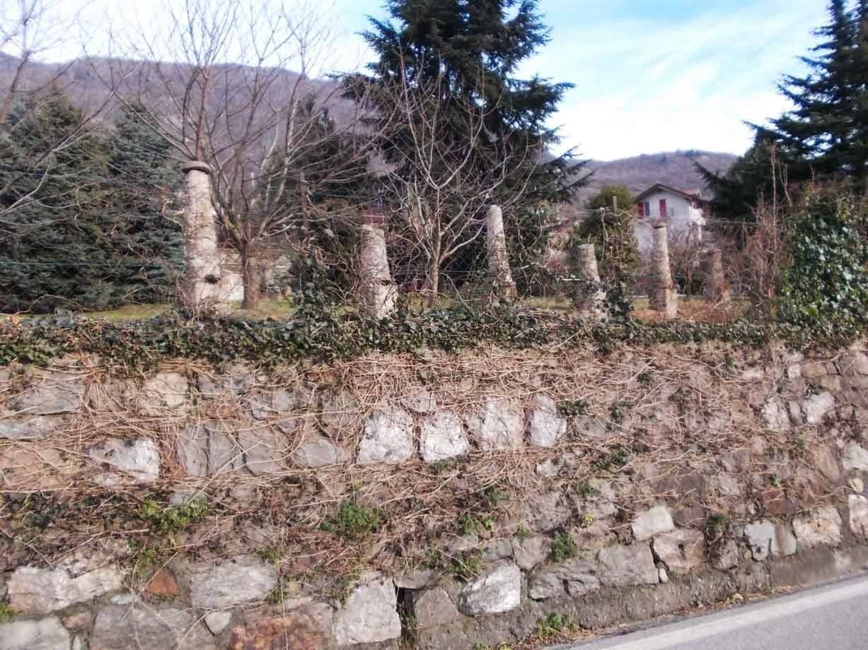 Muro di pietra a secco con piantoni in pietra, edera e conifere, case e monti sullo sfondo