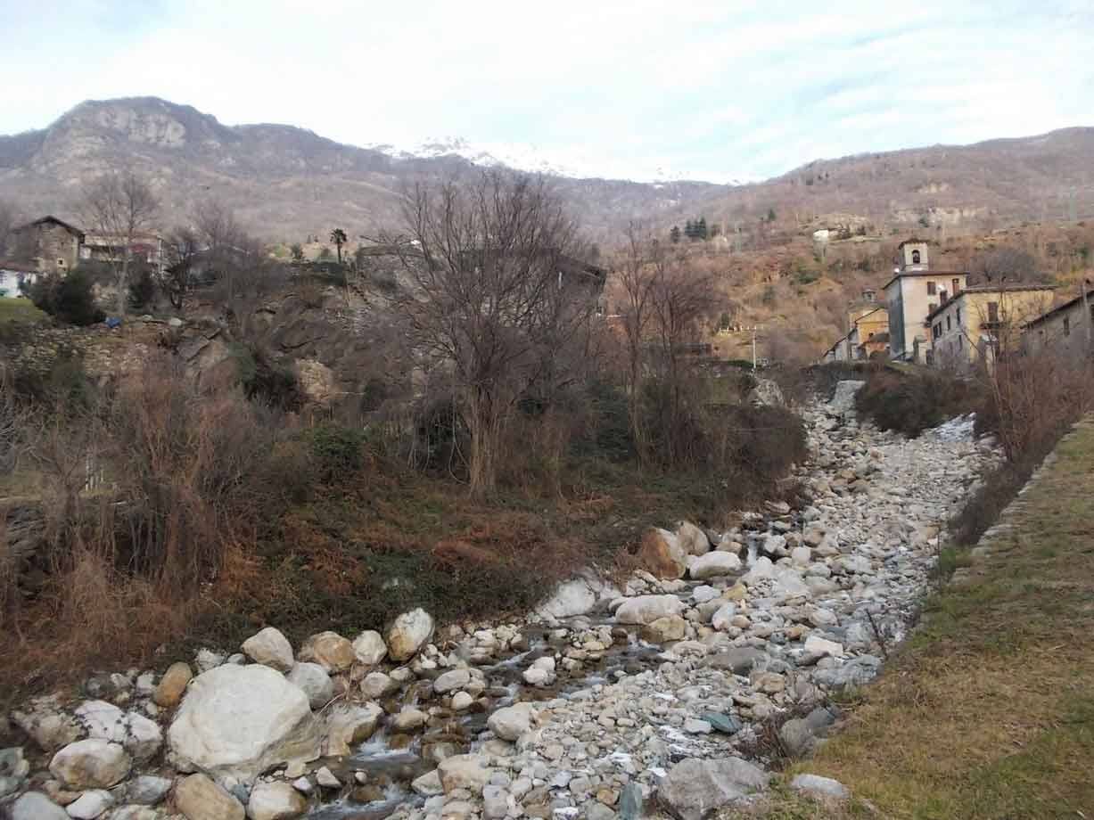 Torrente in secca tra massi, borgo con campanile e monti innevati sullo sfondo