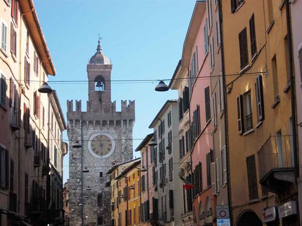 Torre della Pallata in Brescia, stone clock tower rising between colorful buildings and hanging lamps