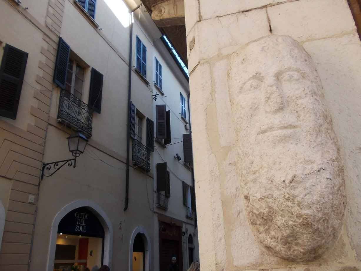 Volto barbuto scolpito nella pietra all'angolo di un vicolo con case e insegna Grotta del Sole
