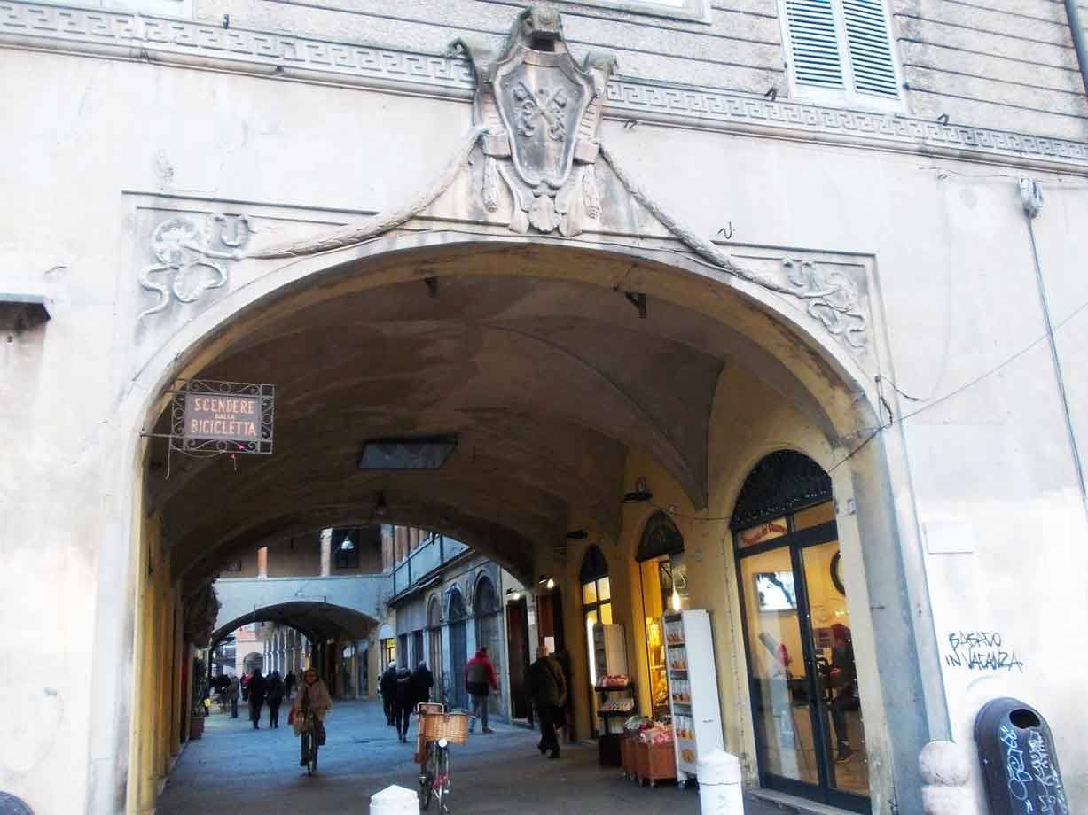 Portico storico con arco decorato e insegna in ferro, passanti e biciclette sotto le volte