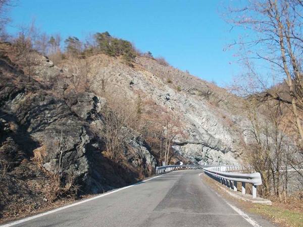 Asphalt road with guardrail between grey rocks and bare trees, blue sky