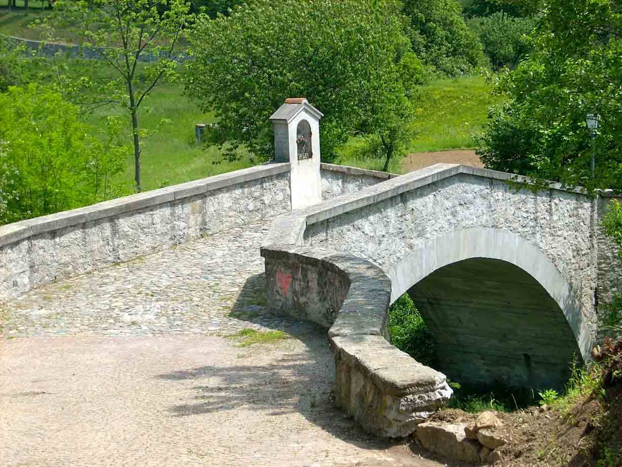 Ponte in pietra ad arco con edicola votiva, prati verdi e alberi illuminati dal sole