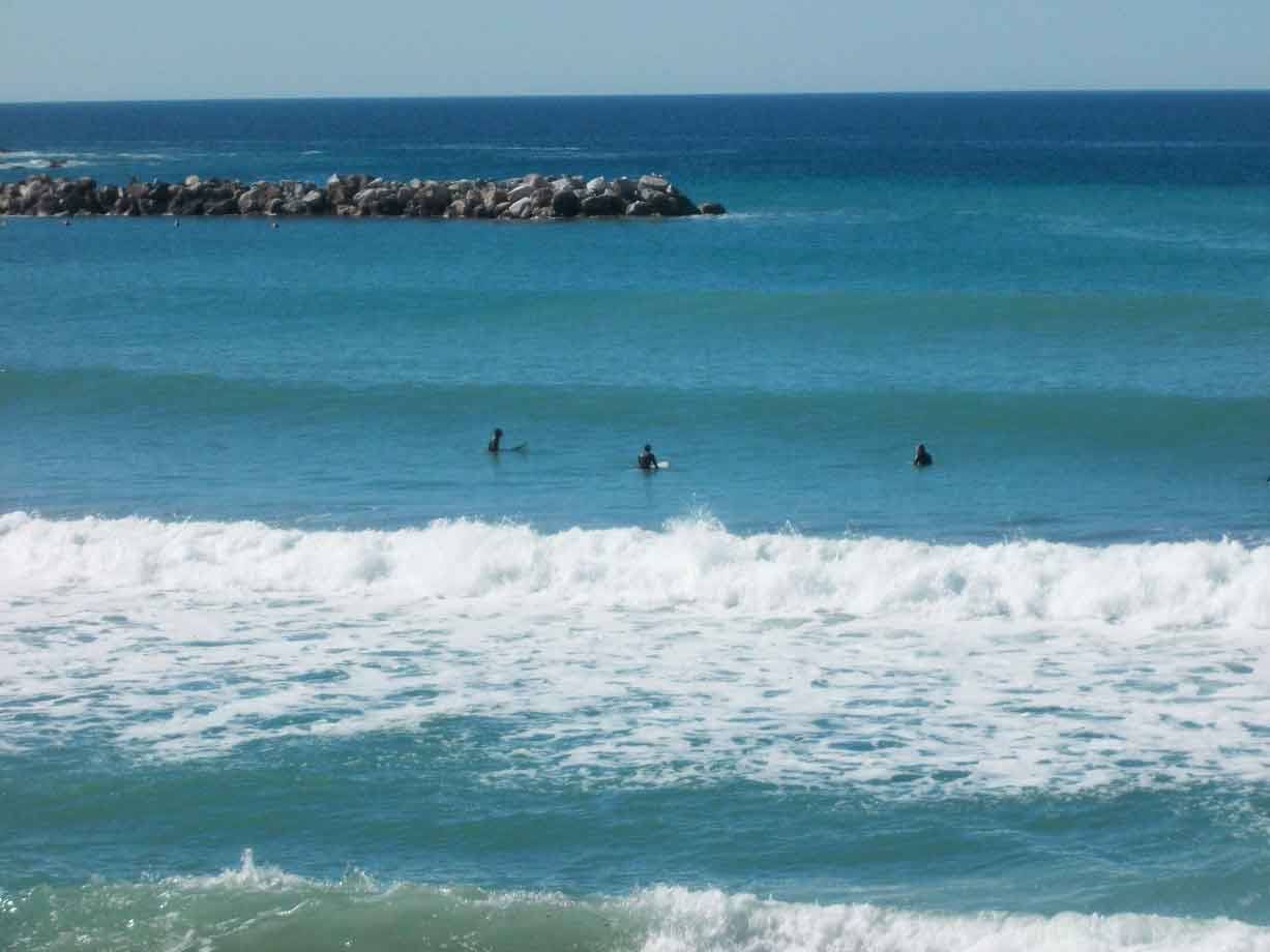 Surfisti in muta aspettano le onde tra la schiuma, scogliera sullo sfondo