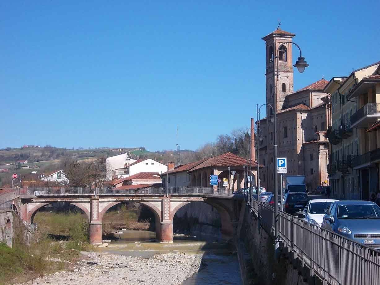Ponte in mattoni sul fiume asciutto, campanile e auto parcheggiate lungo la strada