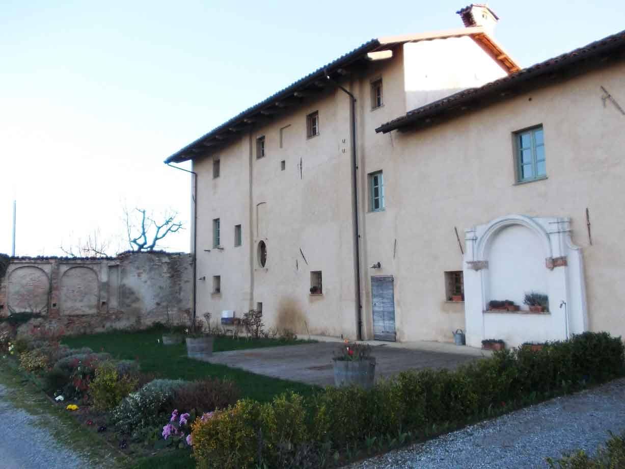 Cascina con facciata intonacata, edicola votiva con vasi di fiori e giardino