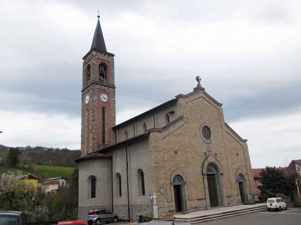 Chiesa in pietra con campanile in mattoni e orellerino, auto parcheggiate sotto il cielo nuvoloso