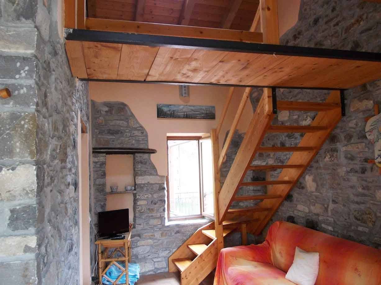 Interno in pietra con scala in legno, soppalco e divano arancione sotto la finestra