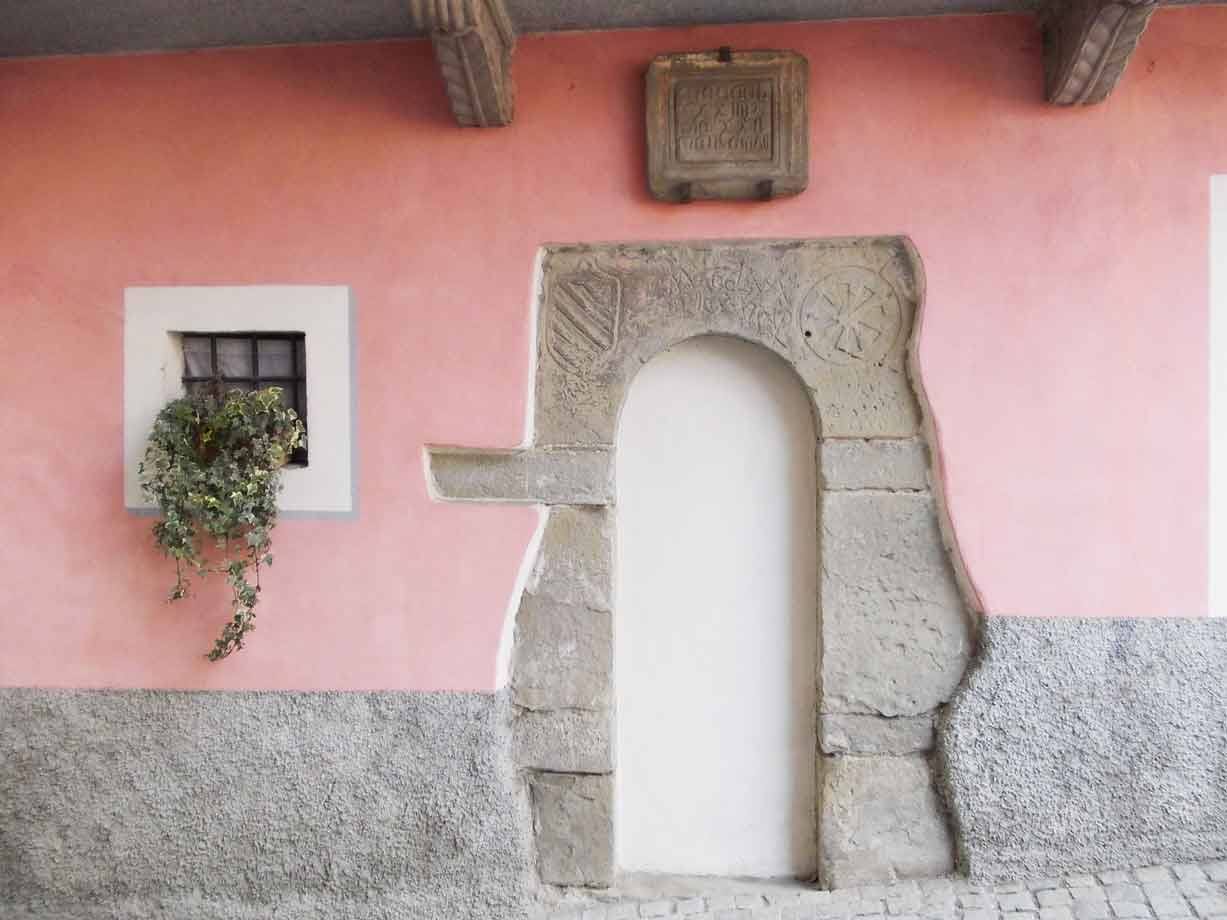 Portale medievale in pietra con stemmi scolpiti, muro rosa, finestra con edera