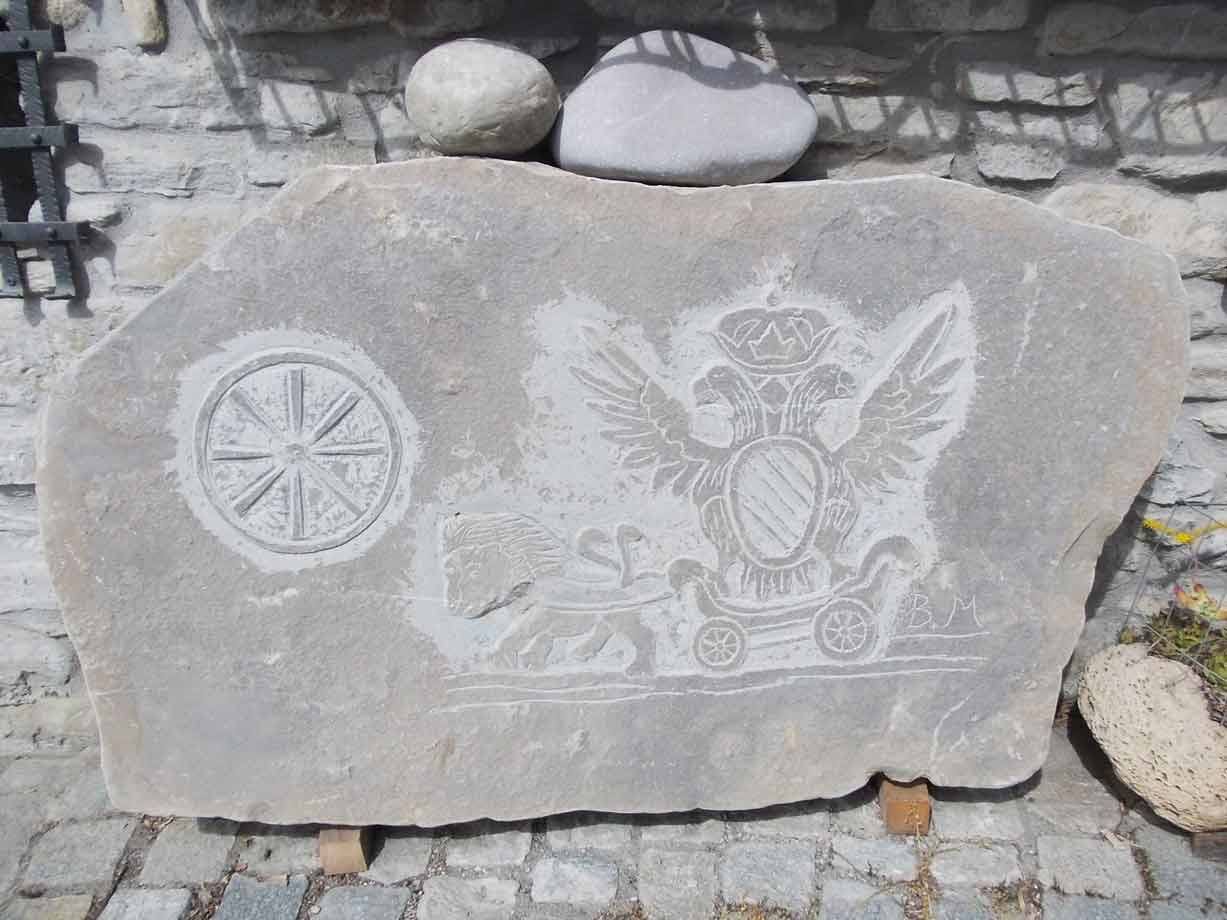 Lastra di pietra incisa con aquila bicipite, corona e ruota, contro muro di pietra