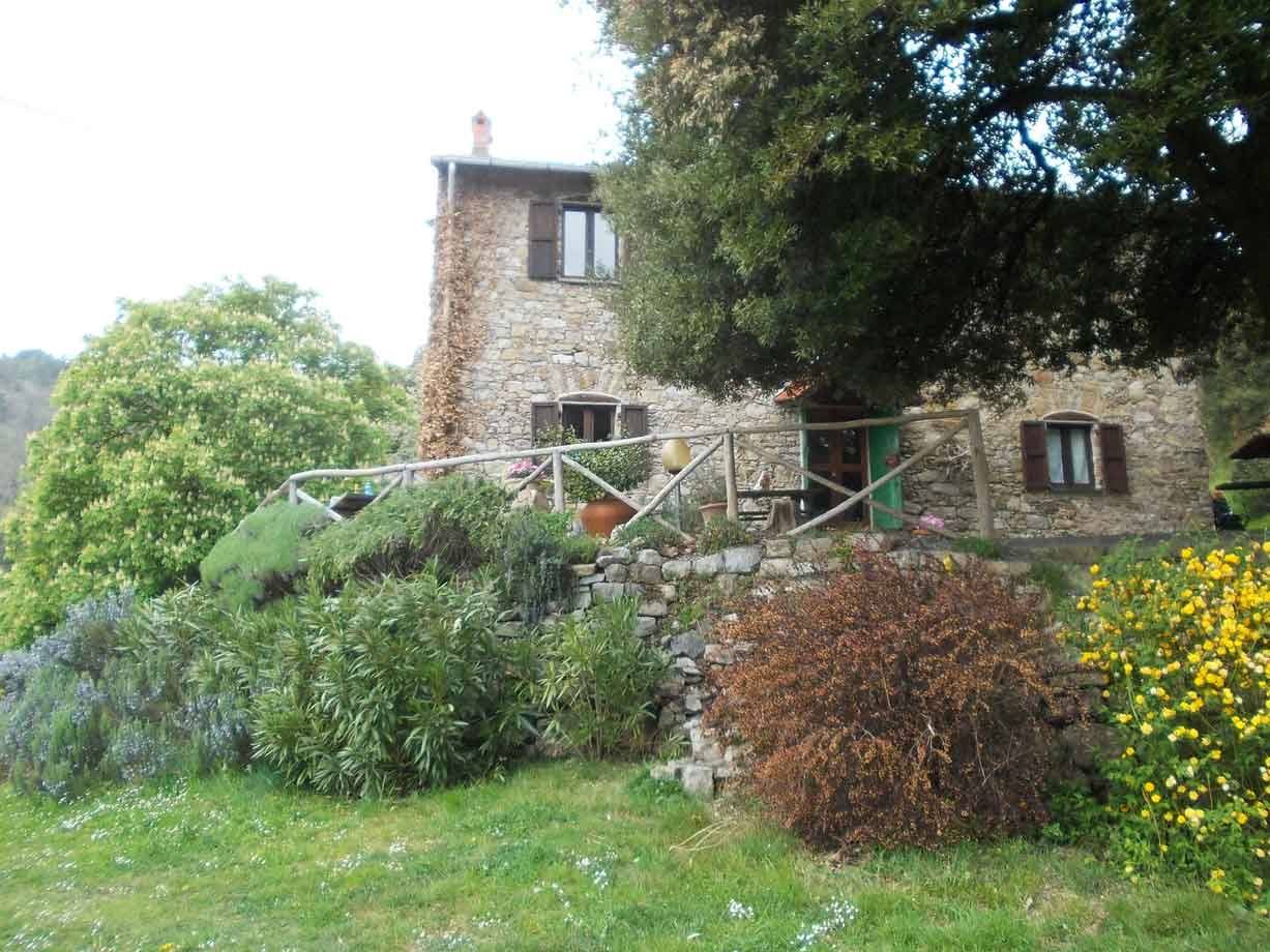 Casa rurale in pietra con persiane verdi, recinzione in legno e giardino fiorito