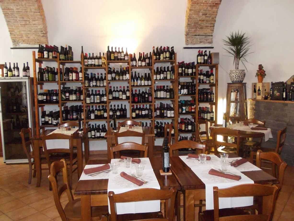 Sala di trattoria con scaffali di legno pieni di bottiglie di vino, tavoli apparecchiati