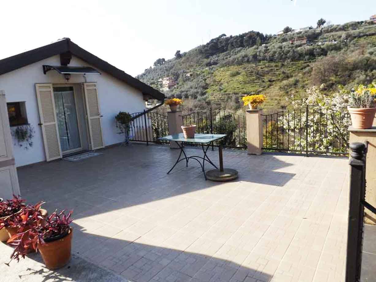 Terrazza piastrellata con tavolo in ferro, vaso di mimosa, colline coltivate dietro la casa
