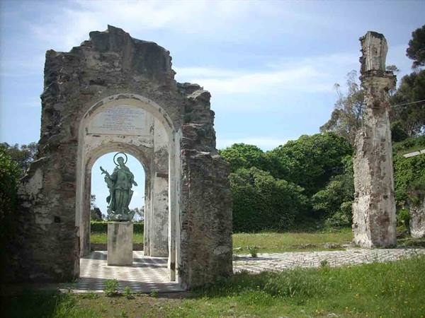 Bronze statue under a stone ruin arch, green trees behind