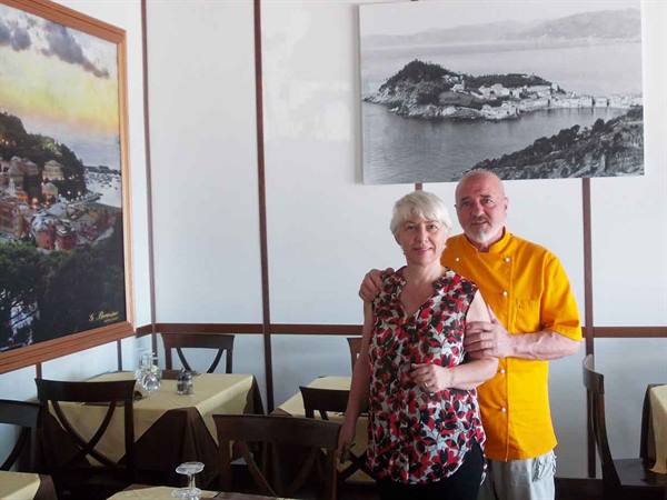 Chef in yellow jacket and woman in restaurant with Sestri Levante paintings