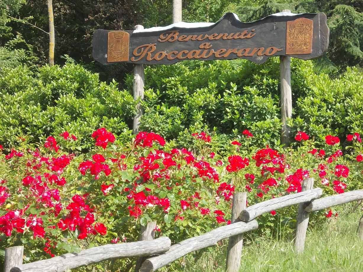 Insegna di benvenuto in legno a Roccaverano tra rose rosse e staccionata