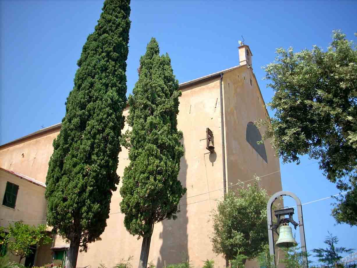 Campanile e campana in ferro davanti a chiesa ocra tra alti cipressi, cielo azzurro