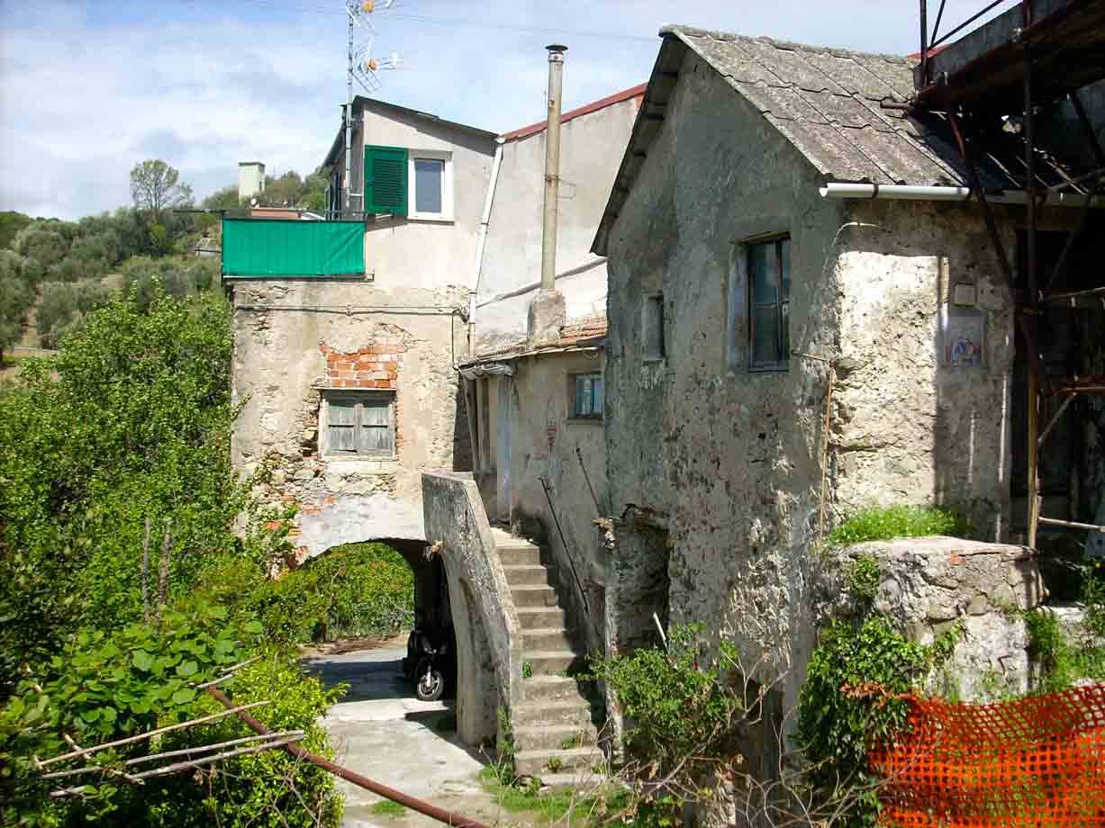 Case rustiche in pietra con scalinata e arco, tappeti verdi appesi, moto all'ombra