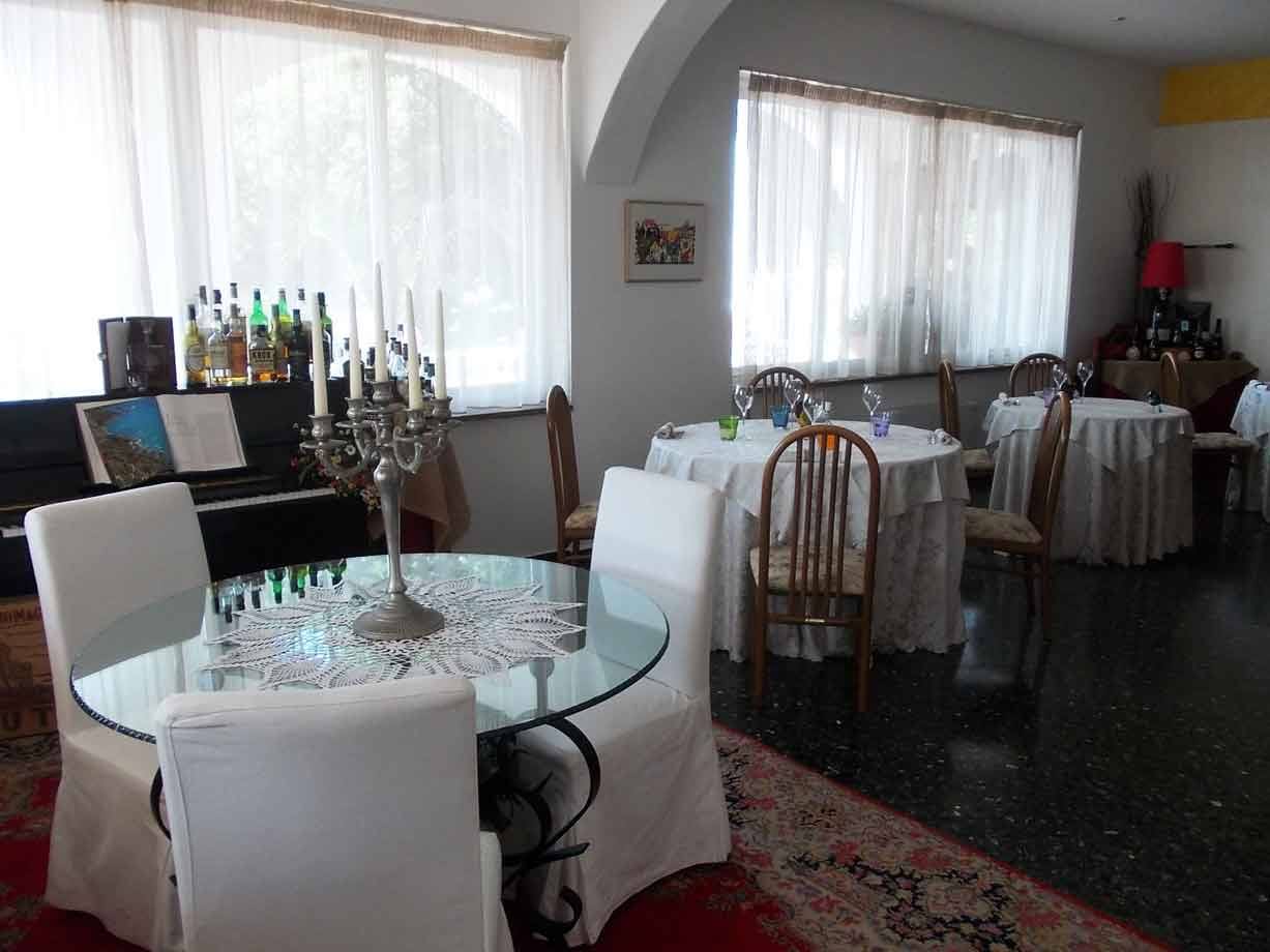 Sala di ristorante con tavoli rotondi in vetro, candele, pianoforte e bottiglie vicino alla finestra