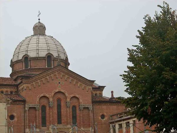 Brick church with grey dome under cloudy sky, green tree beside
