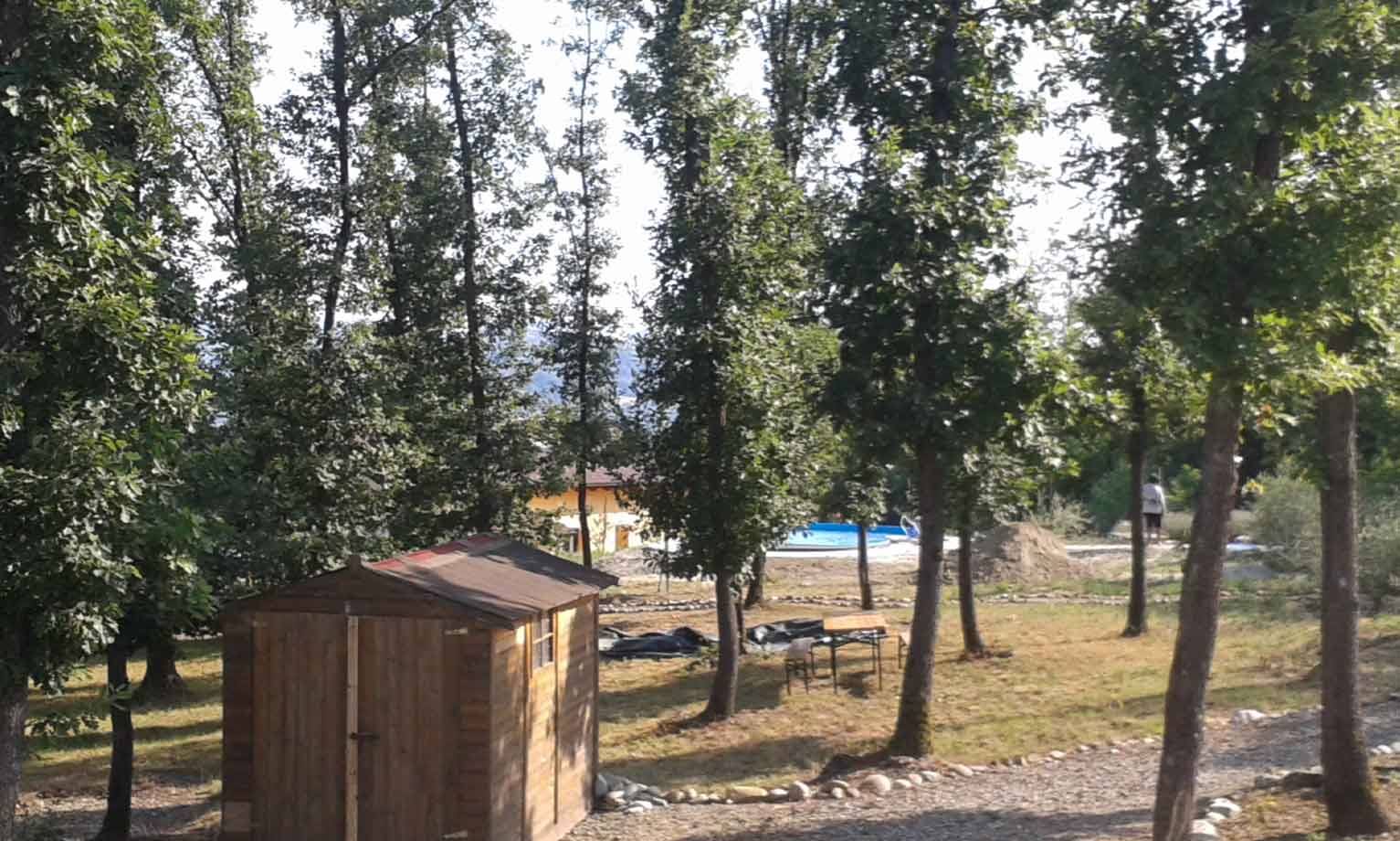 Casetta in legno tra alberi alti, in fondo una piscina e tavoli in erba