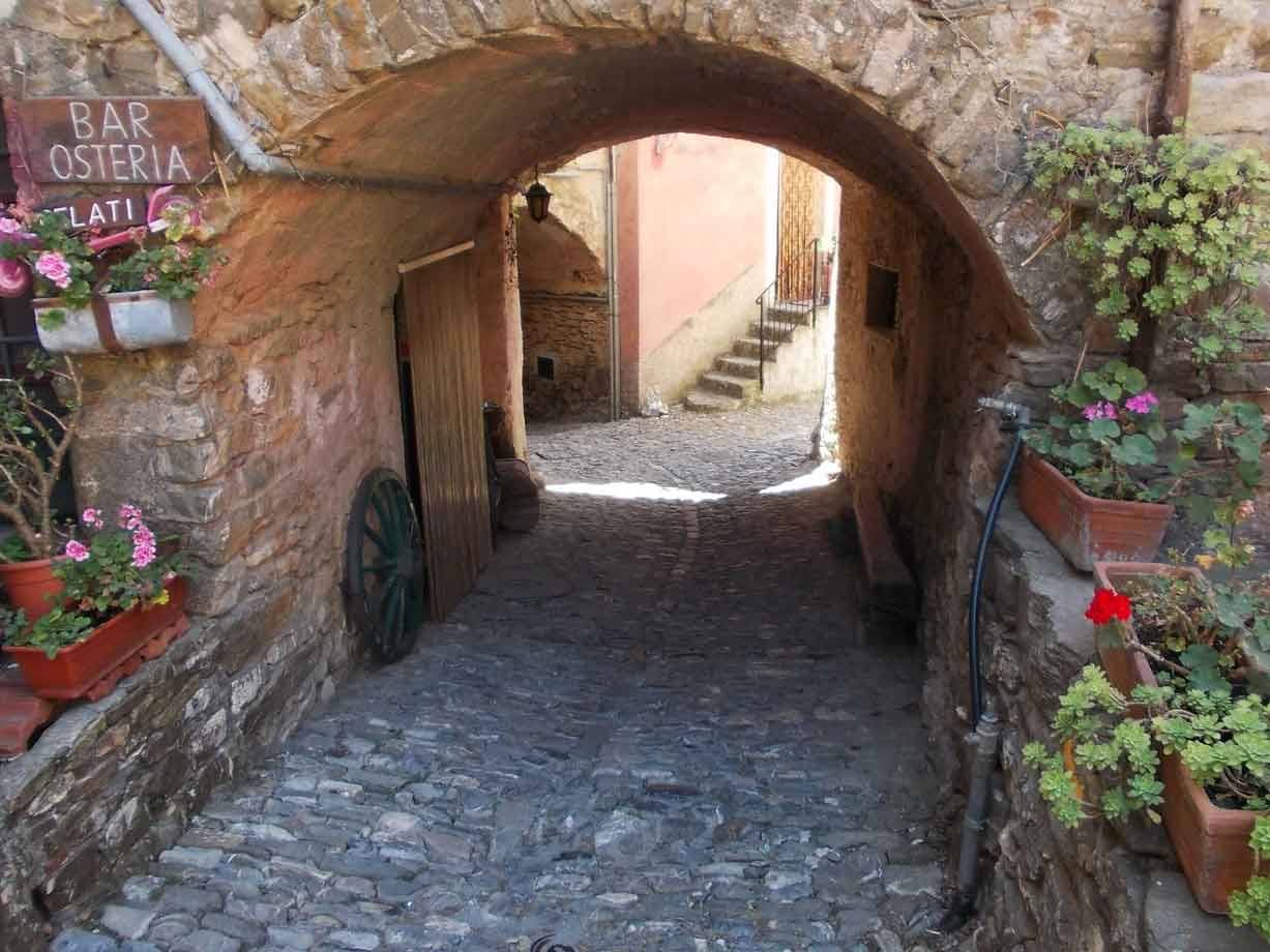 Arco di pietra su vicolo acciottolato, insegna bar osteria e vasi di fiori