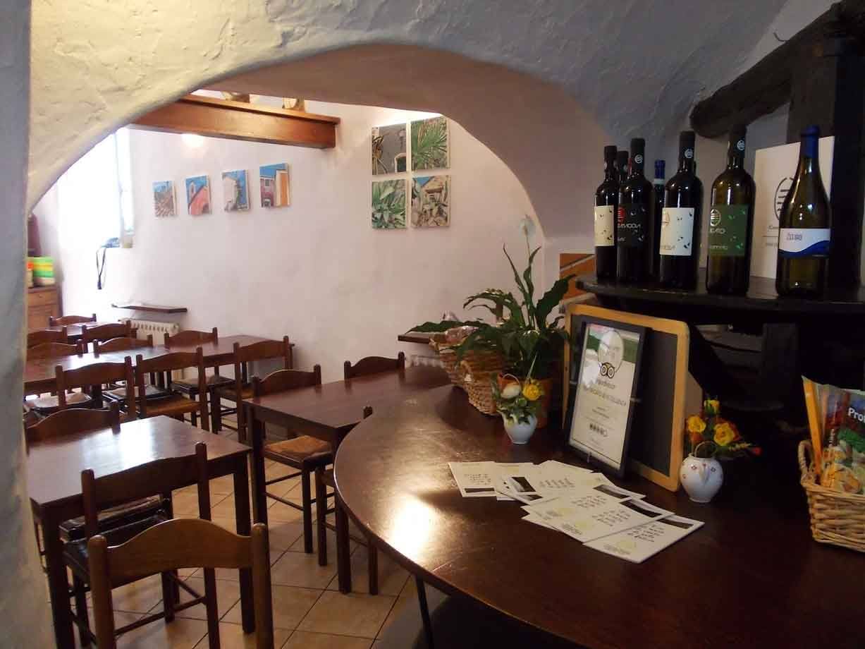 Sala di trattoria con volte bianche, tavoli in legno, bottiglie di vino e quadri