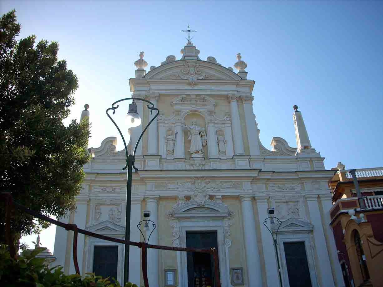 Facciata barocca bianca di chiesa con statue, lampioni e alberi sotto cielo azzurro
