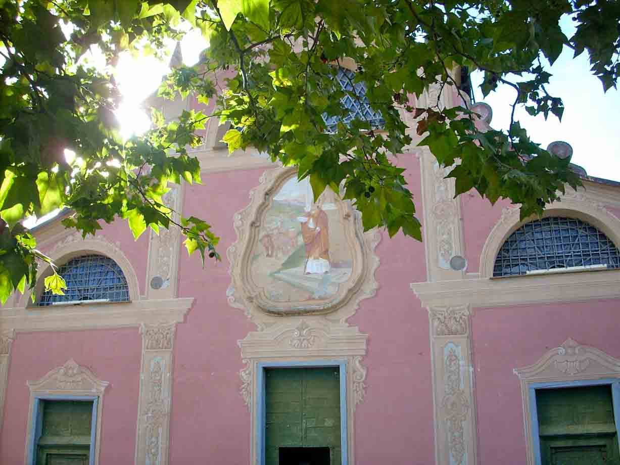 Facciata rosa di chiesa con affresco, finestre e porte verdi tra foglie
