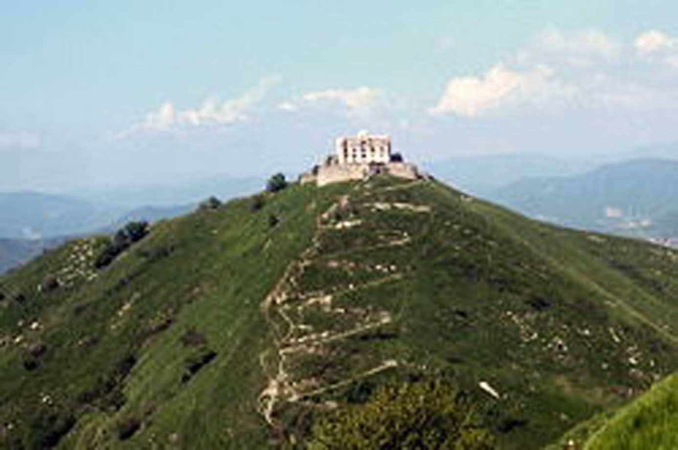 Antico forte in pietra sulla cima verde, sentiero terrazzato sale tra i monti ligure