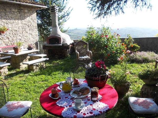 Breakfast on a round table with red cloth, wood oven and roses in a farmhouse garden