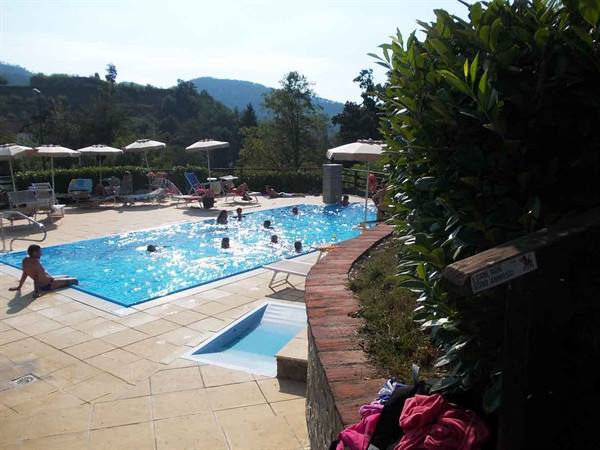 Children swimming in an outdoor pool with umbrellas, loungers and green hills behind