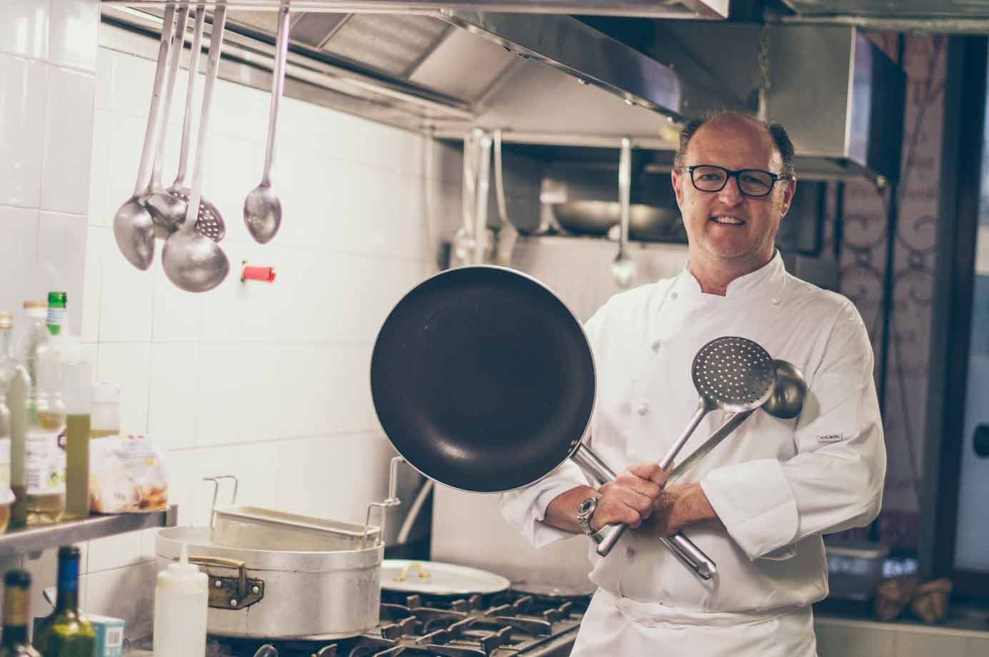 Cuoco sorridente in giacca bianca con padella e mestoli, cucina in acciaio