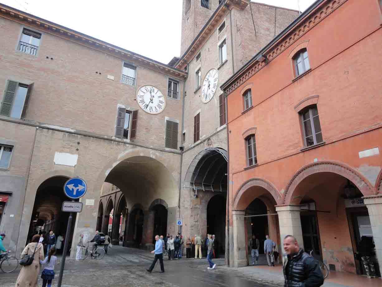 Pedoni sotto i portici dell'Orologio a Bologna, mattoni ocra e cielo grigio