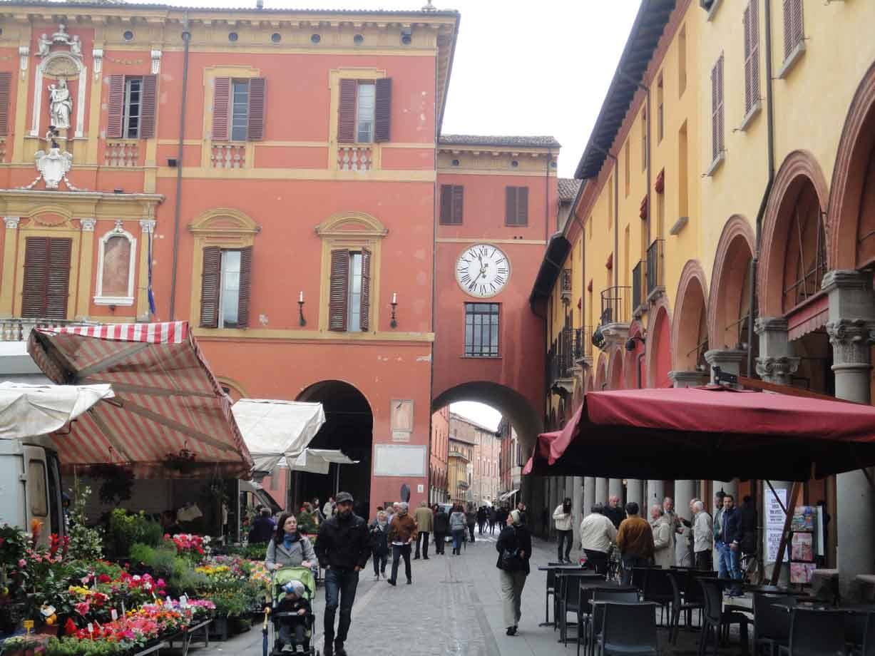 Mercato sotto i portici: bancarella di fiori, orologio sull'arco, passanti