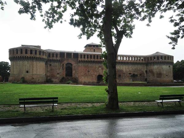 Imola's brick fortress under a grey sky, benches and wet lawn