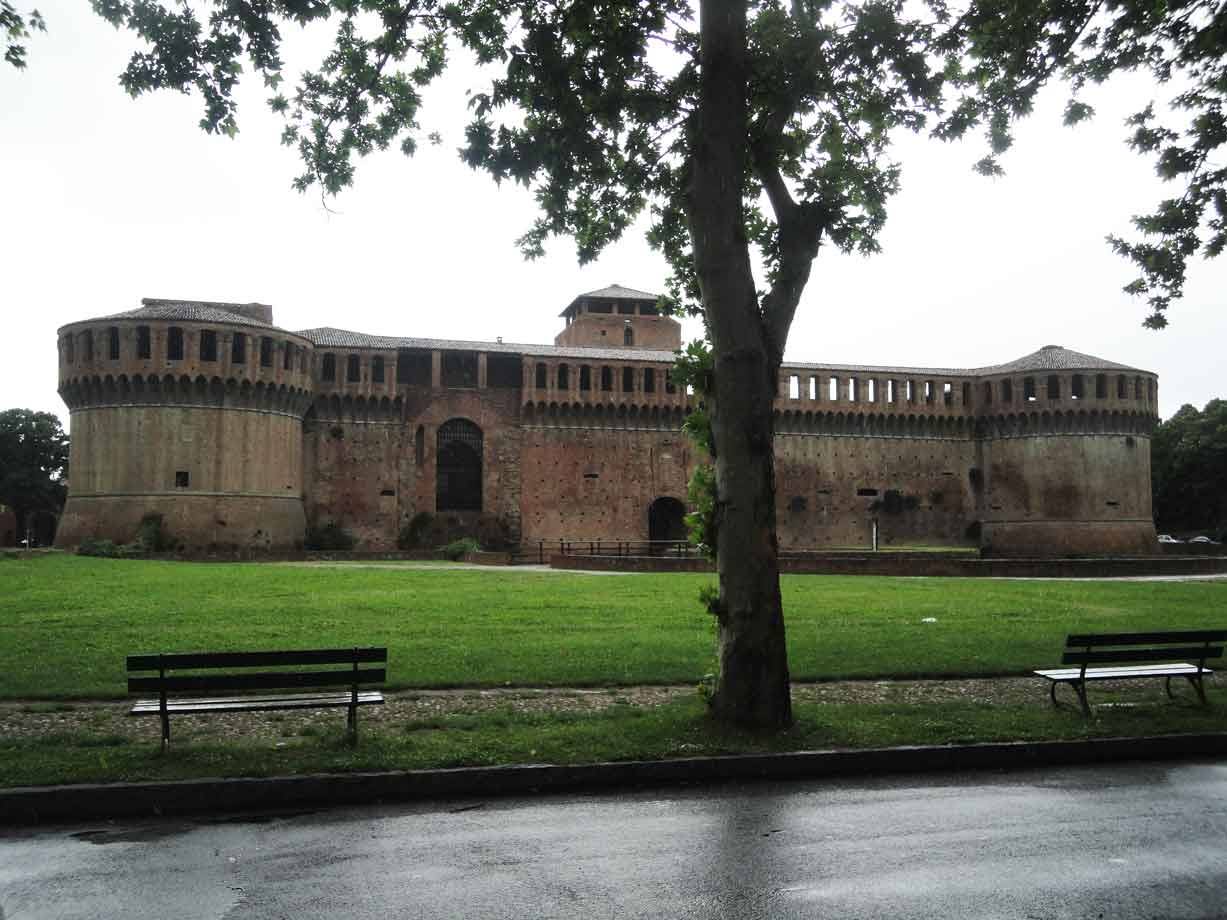 La Rocca di Imola in mattoni sotto il cielo grigio, panchine e prato bagnato