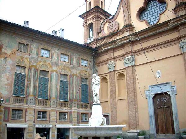 Statue on fountain before a baroque church and faded frescoed palace with green shutters