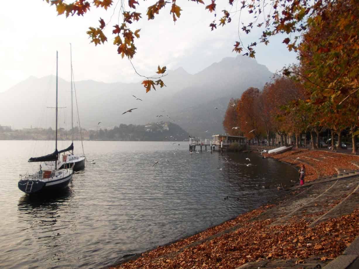 Barca a vela ormeggiata sul lungolago autunnale di Lecco, riva coperta di foglie