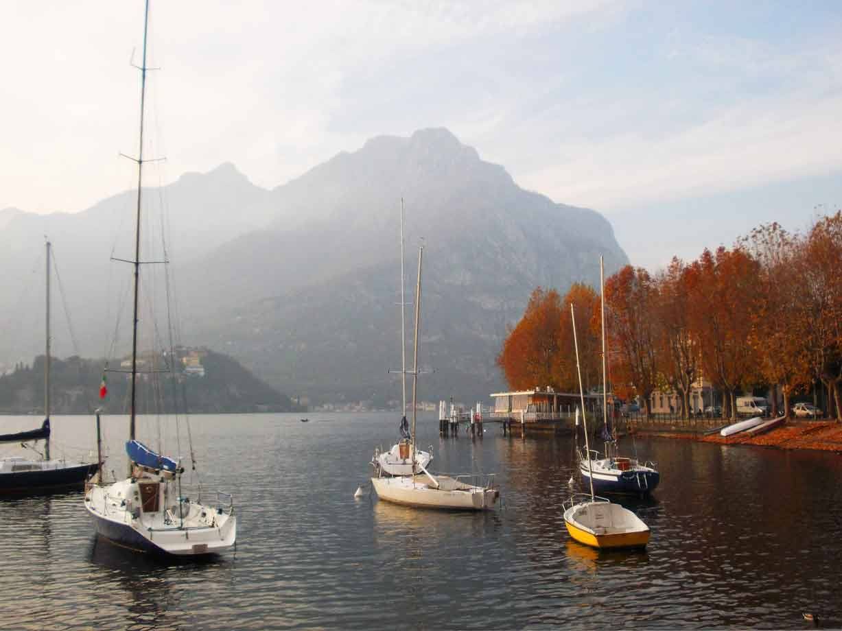 Vele ancorate sul lago di Lecco, monti nebbiosi e alberi autunnali in riva