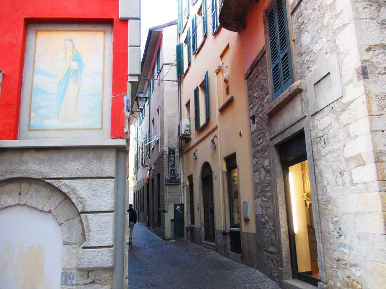 Vicolo stretto tra case con affresco sacro su muro rosso, persiane verdi