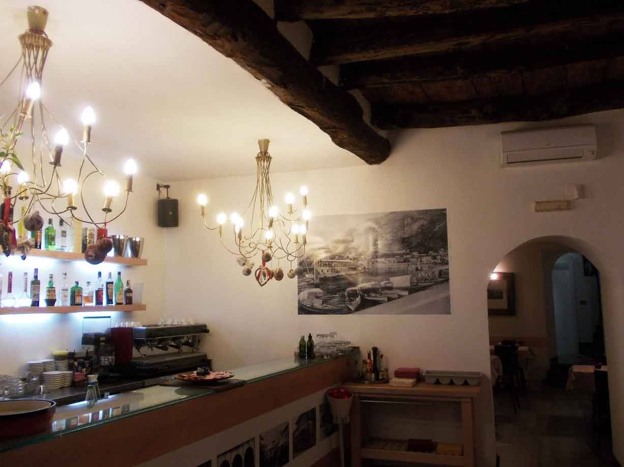 Bar interno con lampadari dorati accesi, bancone, bottiglie e travi lignee