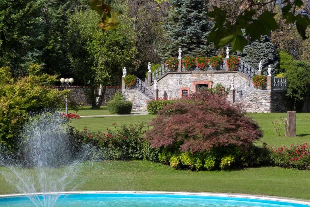 Fontana in primo piano e scalinata in pietra con statue tra giardini fioriti
